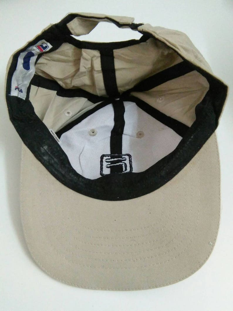May include: A beige baseball cap with a black brim and a black Fila logo on the front. The cap has a black adjustable strap in the back.