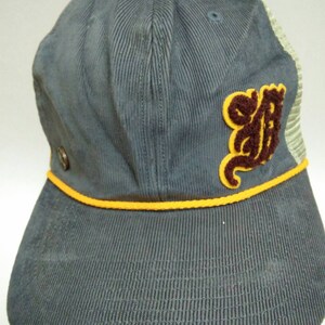 May include: A gray corduroy baseball cap with a yellow trim and a brown and yellow embroidered logo on the back.