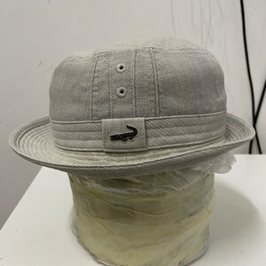 May include: A gray, cotton bucket hat with a small alligator logo on the front. The hat has a wide brim and a slightly rounded crown.