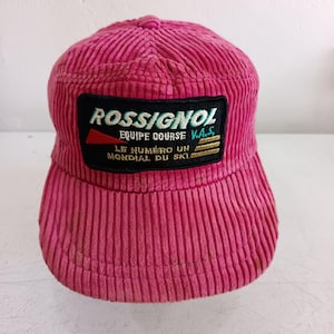 Könnte beinhalten: Eine leuchtend pinke Baseballkappe aus Cord mit einem rechteckigen schwarzen Aufnäher auf der Vorderseite. Der Aufnäher zeigt den weißen Schriftzug ROSSIGNOL, EQUIPE COURSE V.A.S. und LE NUMERO UN MONDIAL DU SKI sowie ein rotes Dreieck und drei goldene Streifen.