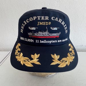 以下が含まれることがあります： ネイビーブルーの野球帽。つばには金色の葉の刺繍が施されています。「HELICOPTER CARRIER」の文字と船のグラフィック、そして「DDH 13,950t 11 helicopters are carried」と書かれています。