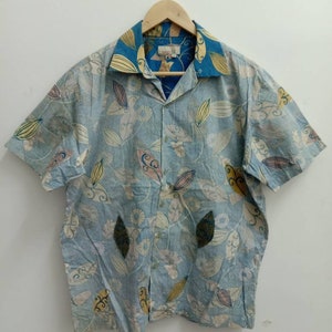 Może przedstawiać: Niebieska koszula z krótkim rękawem zapinana na guziki z kwiatowym wzorem. Koszula ma kołnierzyk z podszewką w niebiesko-żółte wzory.