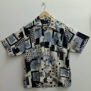 May include: A short-sleeved button-up shirt with a white background and a black, gray, and blue abstract geometric pattern. The shirt has a collar and a button-up front.