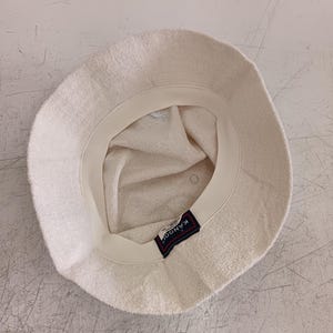 May include: An off-white bucket hat made of a textured terry cloth material. The interior features a cream-colored sweatband and a rectangular black label with the white text KANGOL stitched onto the inner rim of the hat's crown.