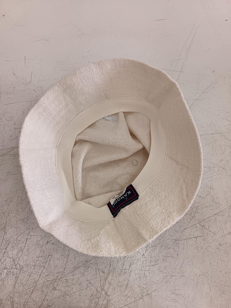 May include: An off-white bucket hat made of a textured terry cloth material. The interior features a cream-colored sweatband and a rectangular black label with the white text KANGOL stitched onto the inner rim of the hat's crown.