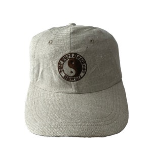 Może przedstawiać: Beżowa czapka z daszkiem z haftowanym brązowym logo z napisem "Tec Surf Dekens Hawaii" i symbolem yin yang.