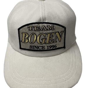 May include: White baseball cap with a black and gold embroidered patch that says "TEAM BOGEN SINCE 1996".