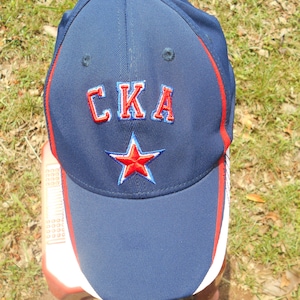 Può includere: Un cappello da baseball blu navy con una stella rossa e il testo "CKA" in lettere rosse e bianche.