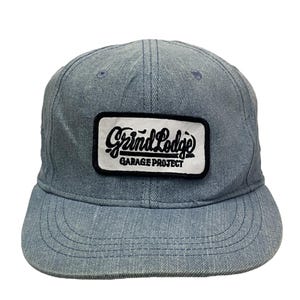 May include: A blue denim baseball cap with a white patch that says "Grind Lodge Garage Project" in black lettering.