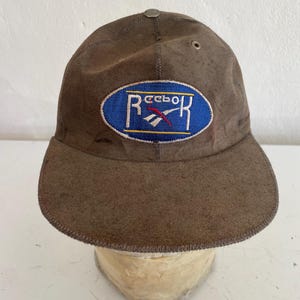 May include: A brown cap with a curved brim. The cap features an embroidered logo in a blue oval with white lettering and a red accent. The cap is made of a worn, textured material and has a small metal button on top.