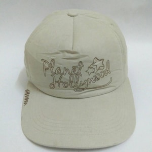May include: A beige baseball cap with the text "Planet Hollywood" embroidered in brown script. The cap features a curved brim and a structured crown. The design includes two stars next to the text.
