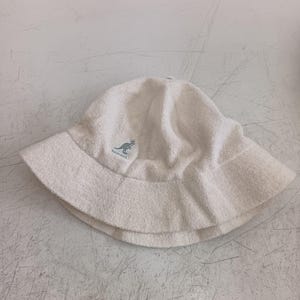 May include: A white bucket hat made of a soft, textured terry cloth material. The hat features a rounded crown and a downward-sloping brim. Embroidered on the front is a small, light blue Kangol logo depicting a kangaroo with the text Kangol.