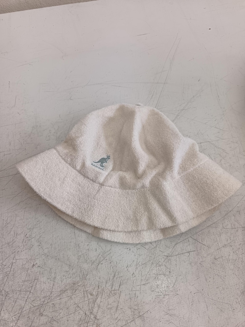 May include: A white bucket hat made of a soft, textured terry cloth material. The hat features a rounded crown and a downward-sloping brim. Embroidered on the front is a small, light blue Kangol logo depicting a kangaroo with the text Kangol.