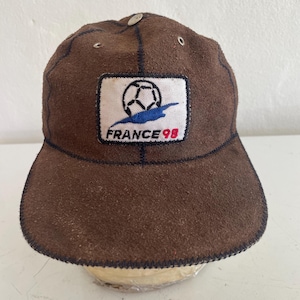 May include: Brown suede baseball cap with a curved brim. The cap features a patch with a soccer ball graphic, a blue swoosh, and the text "FRANCE 98" in red. The cap has a button on top and dark blue stitching.