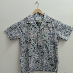 May include: Short-sleeved button-down shirt with a light blue and white geometric pattern featuring various stylized figures. The shirt has a classic collar and a relaxed fit. The label reads "Bai Nani".