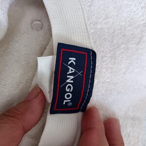 May include: A white textured bucket hat featuring an interior label with a dark blue background, a red border, and the white text KANGOL with a small white kangaroo logo graphic positioned above the letter O on the fabric tag.