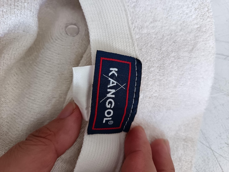 May include: A white textured bucket hat featuring an interior label with a dark blue background, a red border, and the white text KANGOL with a small white kangaroo logo graphic positioned above the letter O on the fabric tag.