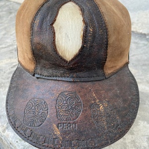May include: A brown leather baseball cap with a white fur patch on the top. The brim of the cap is decorated with a pattern of stylized faces and the word "PERU".