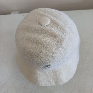 May include: A cream-colored bucket hat with a textured, fuzzy finish. The hat features a small, circular fabric button at the center of the crown and a small embroidered Kangol kangaroo logo in a matching cream tone on the front brim area.