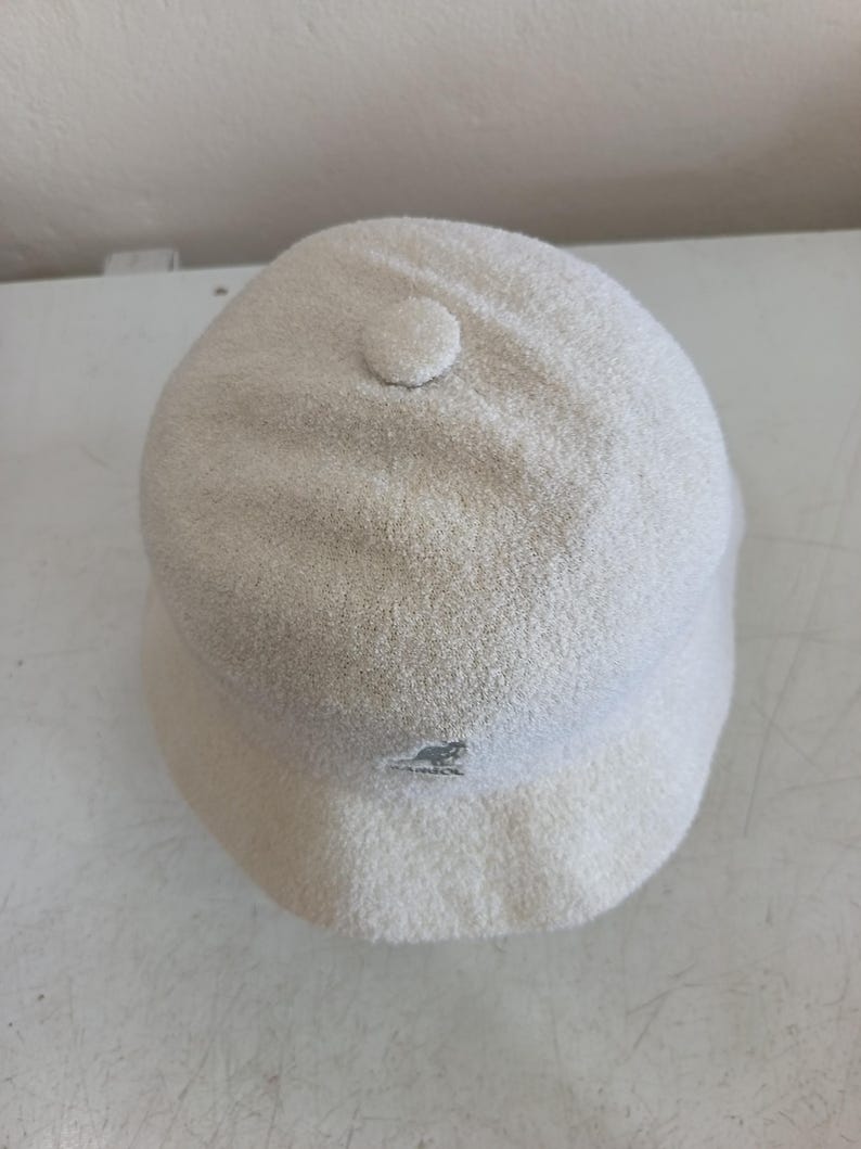 May include: A cream-colored bucket hat with a textured, fuzzy finish. The hat features a small, circular fabric button at the center of the crown and a small embroidered Kangol kangaroo logo in a matching cream tone on the front brim area.