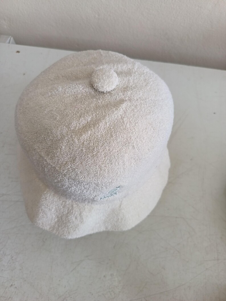 May include: A cream-colored bucket hat with a textured, terry-cloth-like finish. The hat features a small, circular fabric-covered button at the center of the crown and a small embroidered Kangol logo on the side of the brim.