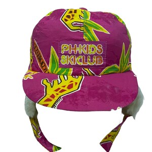 Puede incluir: Sombrero de esquí rosa con borde de piel blanca y estampado de jirafa amarillo y verde. El sombrero tiene un parche en la parte delantera que dice "PH+KIDS SKICLUB".