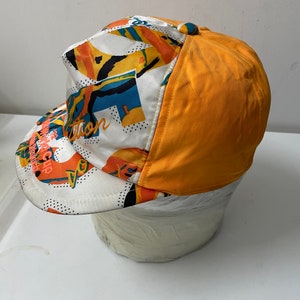 May include: A white and orange baseball cap with a colorful abstract pattern and the text "The Revolution" on the front. The back of the cap is solid orange.