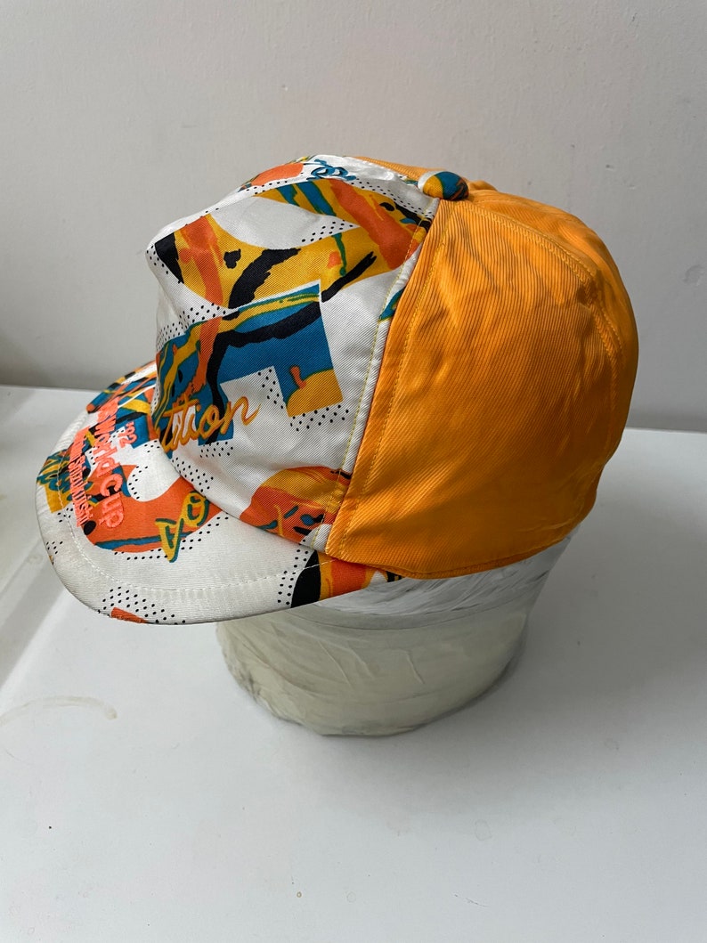 May include: A white and orange baseball cap with a colorful abstract pattern and the text "The Revolution" on the front. The back of the cap is solid orange.