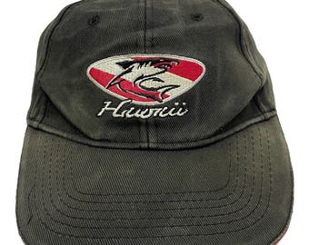 Può includere: Un cappello da baseball verde scuro con un logo di squalo ricamato in rosso, bianco e nero e la parola "Hawaii" sotto.