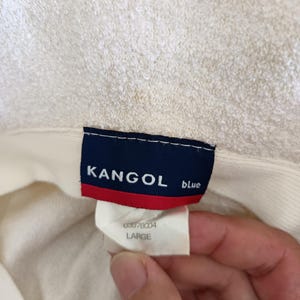 May include: A cream-colored textured hat featuring a rectangular interior label with a dark blue background, white text reading KANGOL blue, and a red horizontal stripe. A secondary white tag below displays the text 03978004 and LARGE.