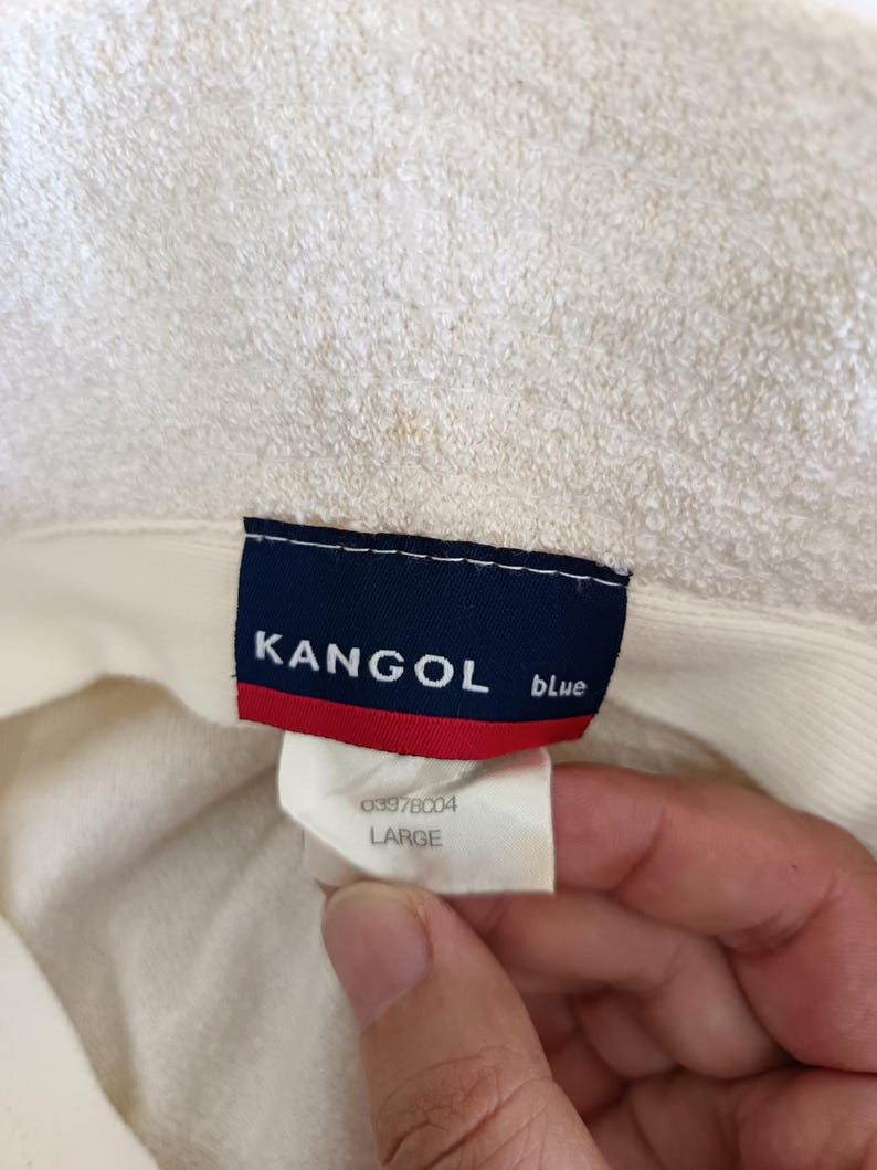 May include: A cream-colored textured hat featuring a rectangular interior label with a dark blue background, white text reading KANGOL blue, and a red horizontal stripe. A secondary white tag below displays the text 03978004 and LARGE.
