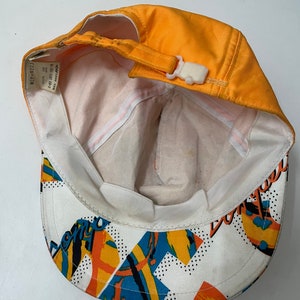 May include: Orange and white baseball cap with a colorful geometric pattern and the word "Don't" on the back. The cap has a white adjustable strap with a plastic buckle.