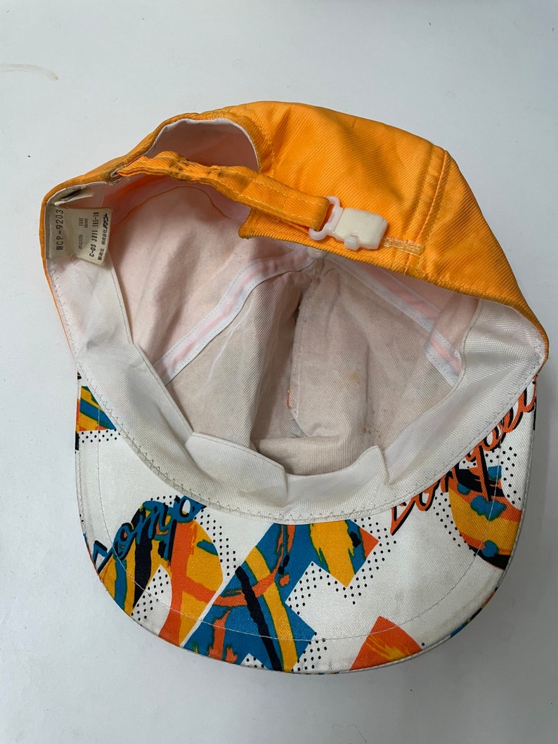 May include: Orange and white baseball cap with a colorful geometric pattern and the word "Don't" on the back. The cap has a white adjustable strap with a plastic buckle.