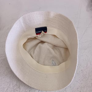 May include: An off-white bucket hat with a textured fabric finish. The interior features a cream-colored sweatband and a dark blue rectangular tag with the white text KANGOL. A small, light-colored embroidered kangaroo logo is visible on the inner crown.