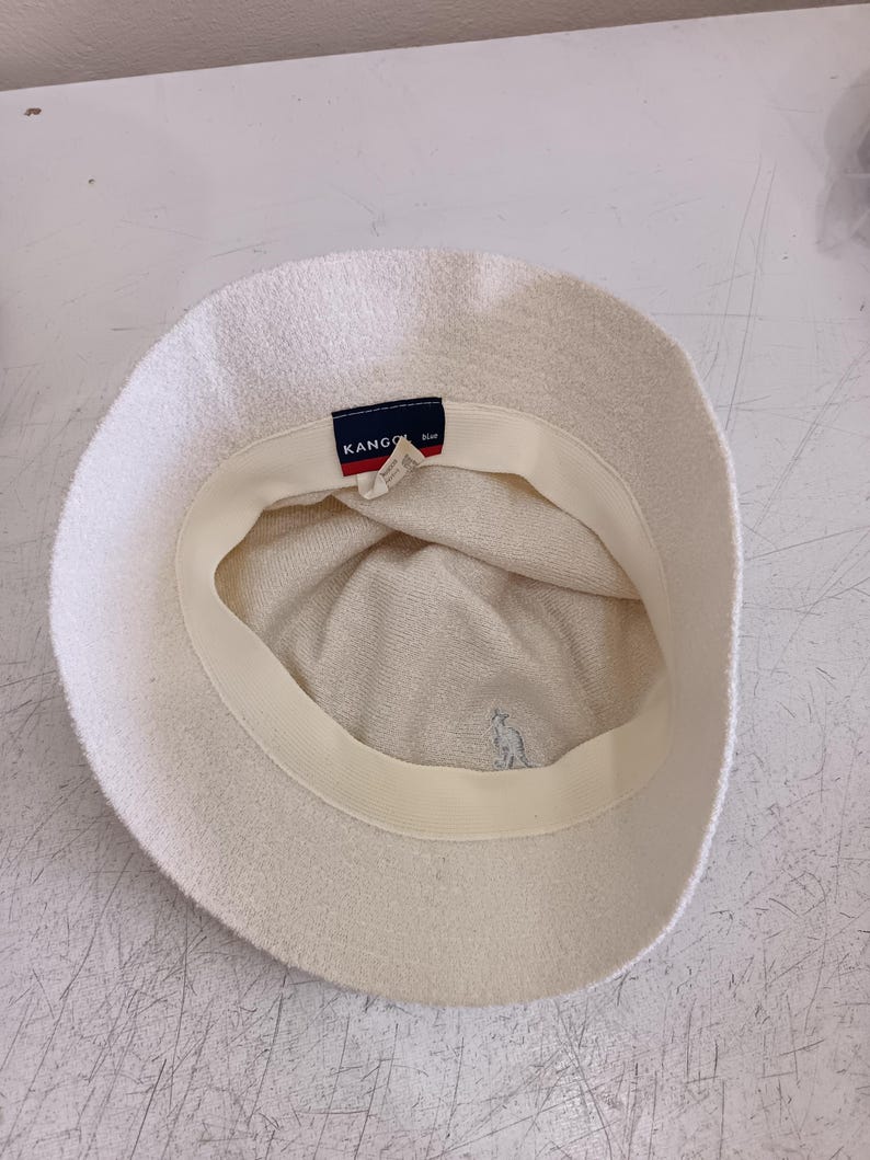 May include: An off-white bucket hat with a textured fabric finish. The interior features a cream-colored sweatband and a dark blue rectangular tag with the white text KANGOL. A small, light-colored embroidered kangaroo logo is visible on the inner crown.