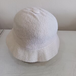 May include: A solid off-white bucket hat with a rounded crown and a short, downward-sloping brim. The hat is crafted from a textured, terry cloth-like fabric that features a soft, looped pile surface throughout the entire exterior of the headwear.