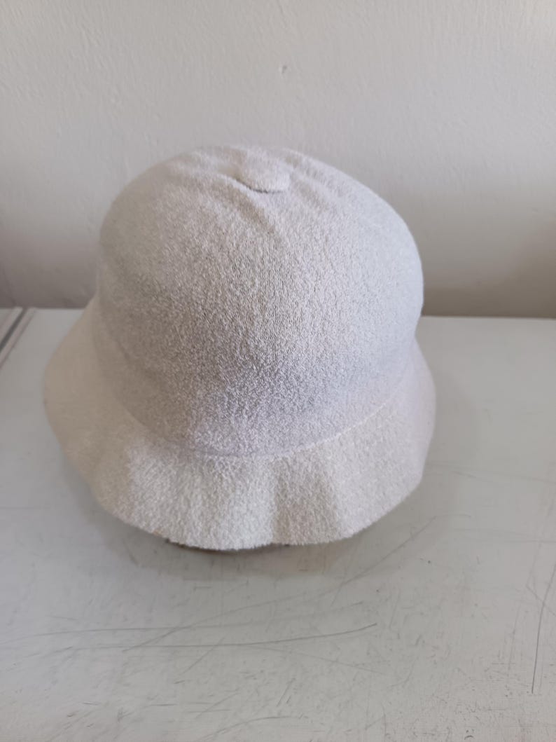 May include: A solid off-white bucket hat with a rounded crown and a short, downward-sloping brim. The hat is crafted from a textured, terry cloth-like fabric that features a soft, looped pile surface throughout the entire exterior of the headwear.