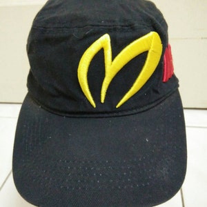 Puede incluir: Una gorra de béisbol negra con una letra 'M' bordada en amarillo en la parte delantera. La gorra tiene un logotipo rojo y amarillo en el lateral.