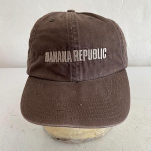 May include: A brown baseball cap with the text "BANANA REPUBLIC" embroidered in white across the front. The cap has a curved brim and a fabric strap on the back for adjustment. The hat is made of a durable, cotton-like material.