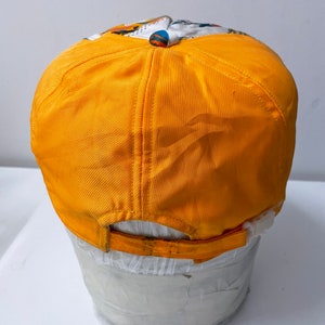 May include: A yellow baseball cap with a white and colorful patterned design on the front. The cap is sitting on a white mannequin head.