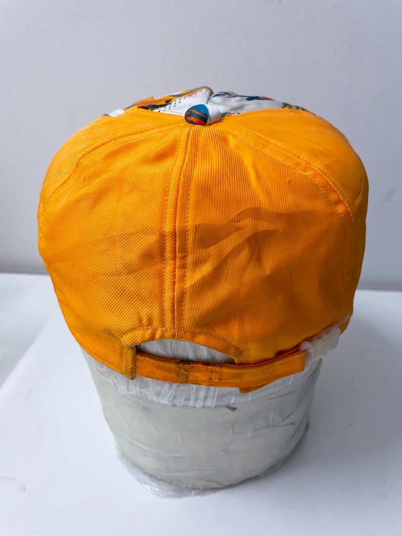 May include: A yellow baseball cap with a white and colorful patterned design on the front. The cap is sitting on a white mannequin head.