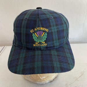 May include: A navy blue and green plaid baseball cap with the text "ST. ANDREWS SCOTLAND" embroidered in gold and green. The cap features a thistle and golf club emblem.