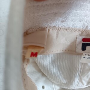 Pode incluir: O interior de um chap&eacute;u branco com uma faixa de suor bege e uma etiqueta de tamanho M em vermelho. Uma etiqueta de tecido branco exibe o logotipo da Fila em vermelho e azul marinho, junto com o texto FL 9700. O interior &eacute; de malha branca.