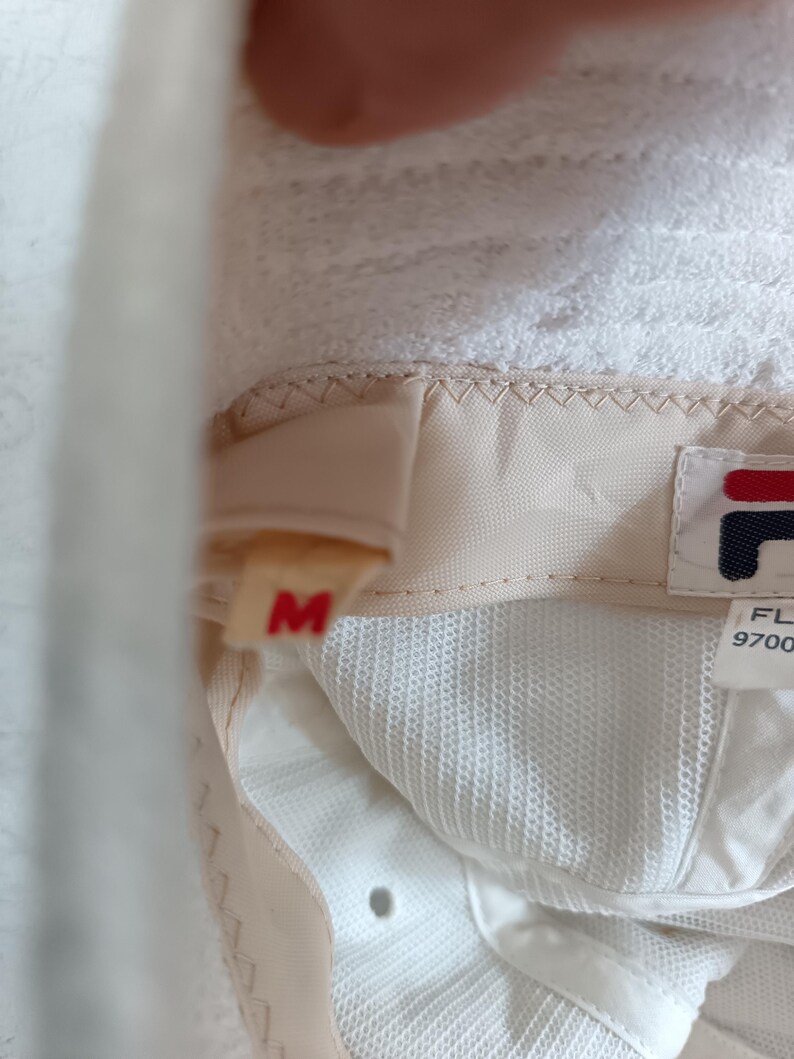 Pode incluir: O interior de um chap&eacute;u branco com uma faixa de suor bege e uma etiqueta de tamanho M em vermelho. Uma etiqueta de tecido branco exibe o logotipo da Fila em vermelho e azul marinho, junto com o texto FL 9700. O interior &eacute; de malha branca.