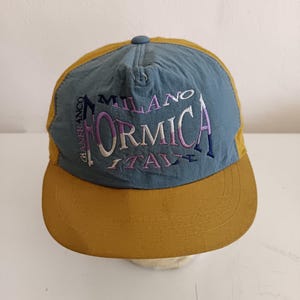 May include: A baseball cap with a blue crown and a mustard yellow brim. The cap features embroidered text that reads "MILANO FORMICA ITALY" in purple and white. The brand name "GIANFRANCO" is also embroidered on the side.