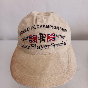 May include: A beige cap with embroidered text and logos. The text reads "WORLD F-1 CHAMPIONSHIP TEAM LOTUS John Player Special" with two Union Jack flags. The cap has a curved brim and a soft, textured appearance.