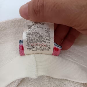 May include: A white fabric care label attached to a textured off-white garment. The label displays the number 141369003, material composition percentages, Japanese care instructions, laundry symbols, and the text Kurihara Corporation Tokyo Made in China.