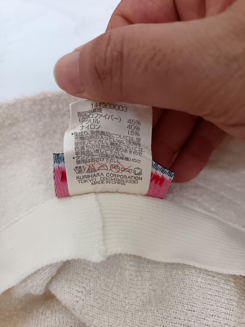 May include: A white fabric care label attached to a textured off-white garment. The label displays the number 141369003, material composition percentages, Japanese care instructions, laundry symbols, and the text Kurihara Corporation Tokyo Made in China.