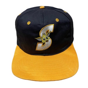 May include: A black and gold baseball cap with a cartoon character wearing a white 'S' on the front. The character has yellow fur and a white face.