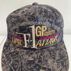 以下が含まれることがあります： 色あせた黒とグレーの模様のデニムスタイルのキャップ。キャップには、「WORLD CHAMPIONSHIP ROUND FOR F1 PILOTS」と「F1 GP ATTACK SAN MARINO MONACO」の刺繍が金、赤、白で施されています。